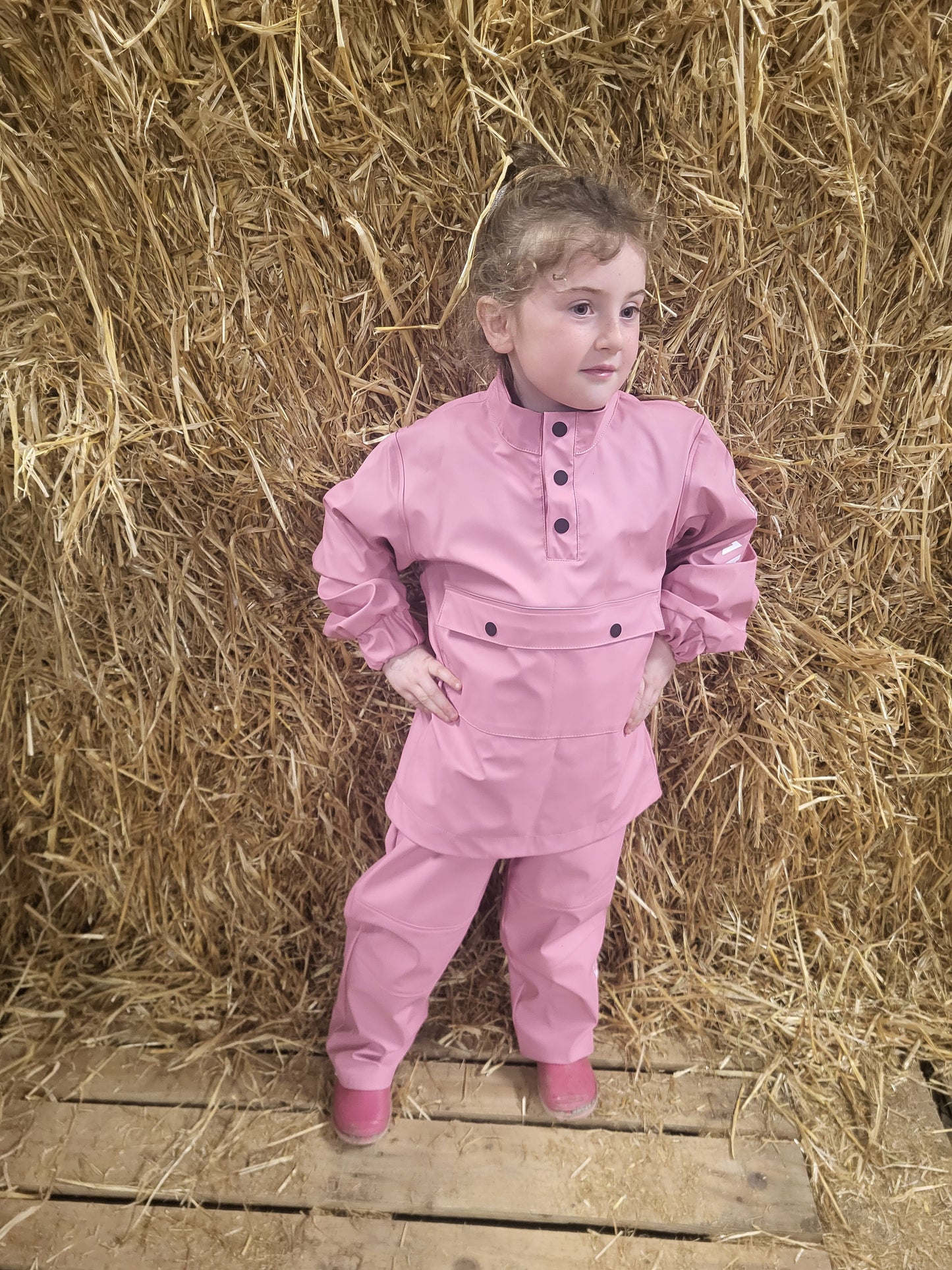 A child standing on a wooden platform by a stack of hay, wearing a Wawets pink waterproof top and pants set"