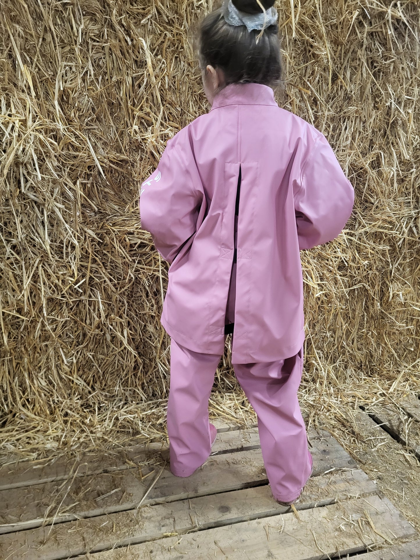 A child standing on a wooden platform by a stack of hay, wearing a wawets waterproof pinktop with velcro back and pink waterproof elastic pants