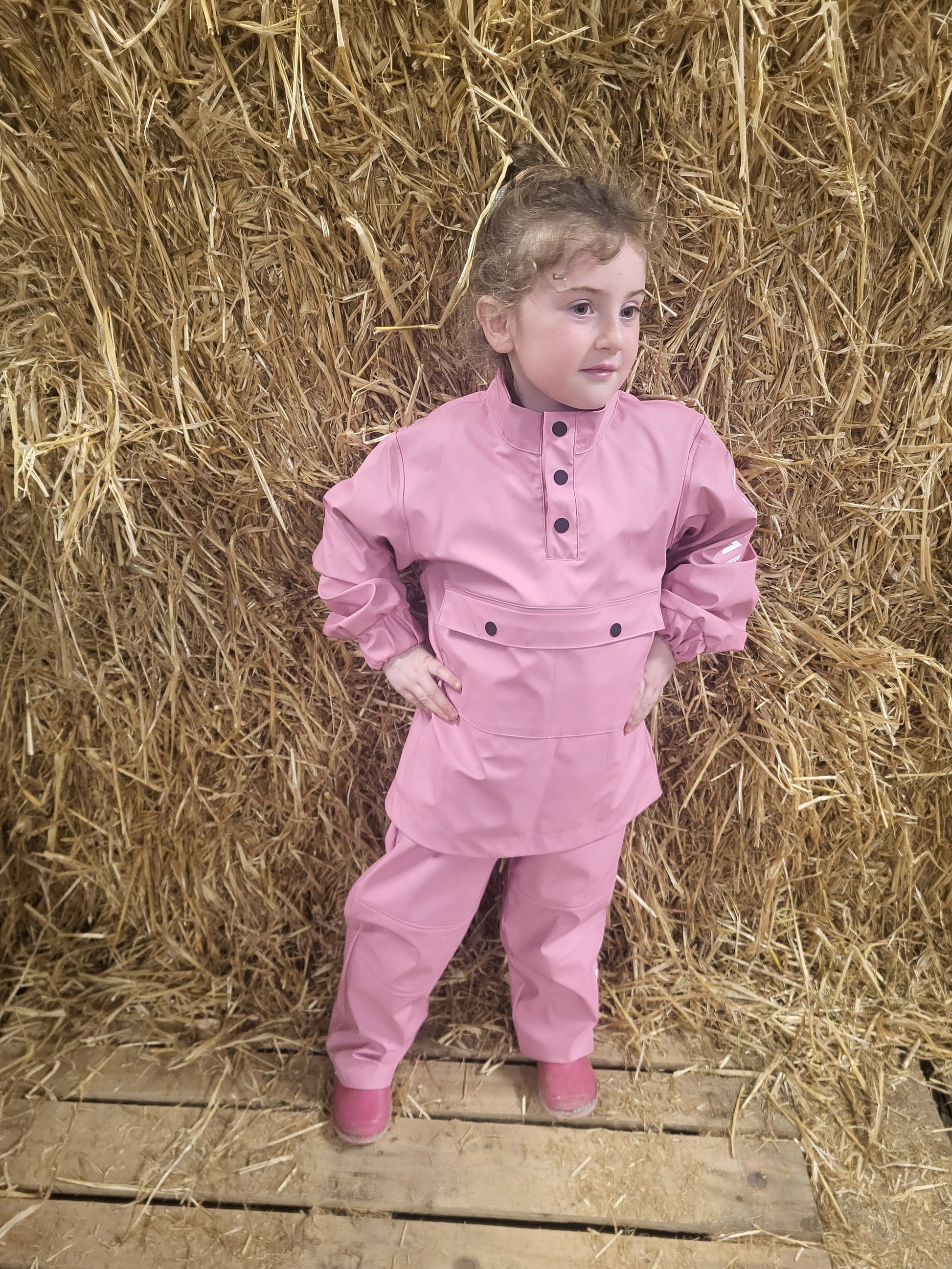 A child standing on a wooden platform by a stack of hay, wearing a Wawets pink waterproof top and pants set"