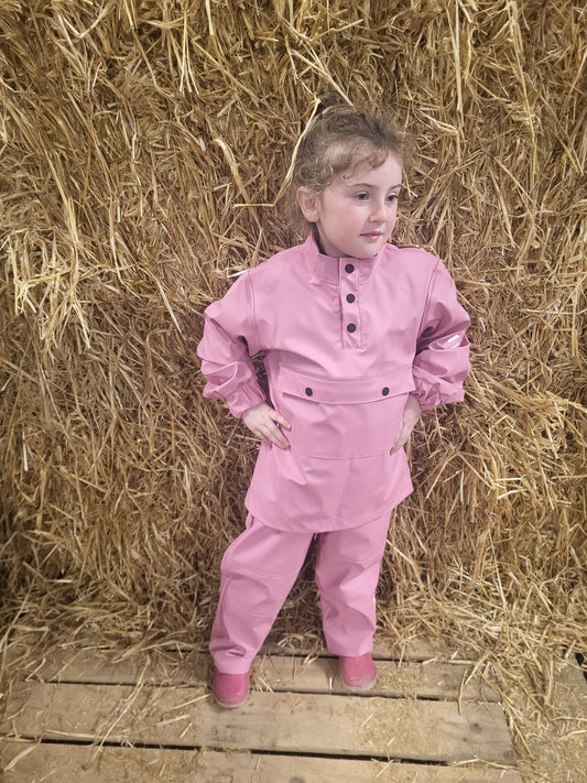 A child standing on a wooden platform by a stack of hay, wearing a pink wawets waterproof top and elastic waist pants with button close at ankle